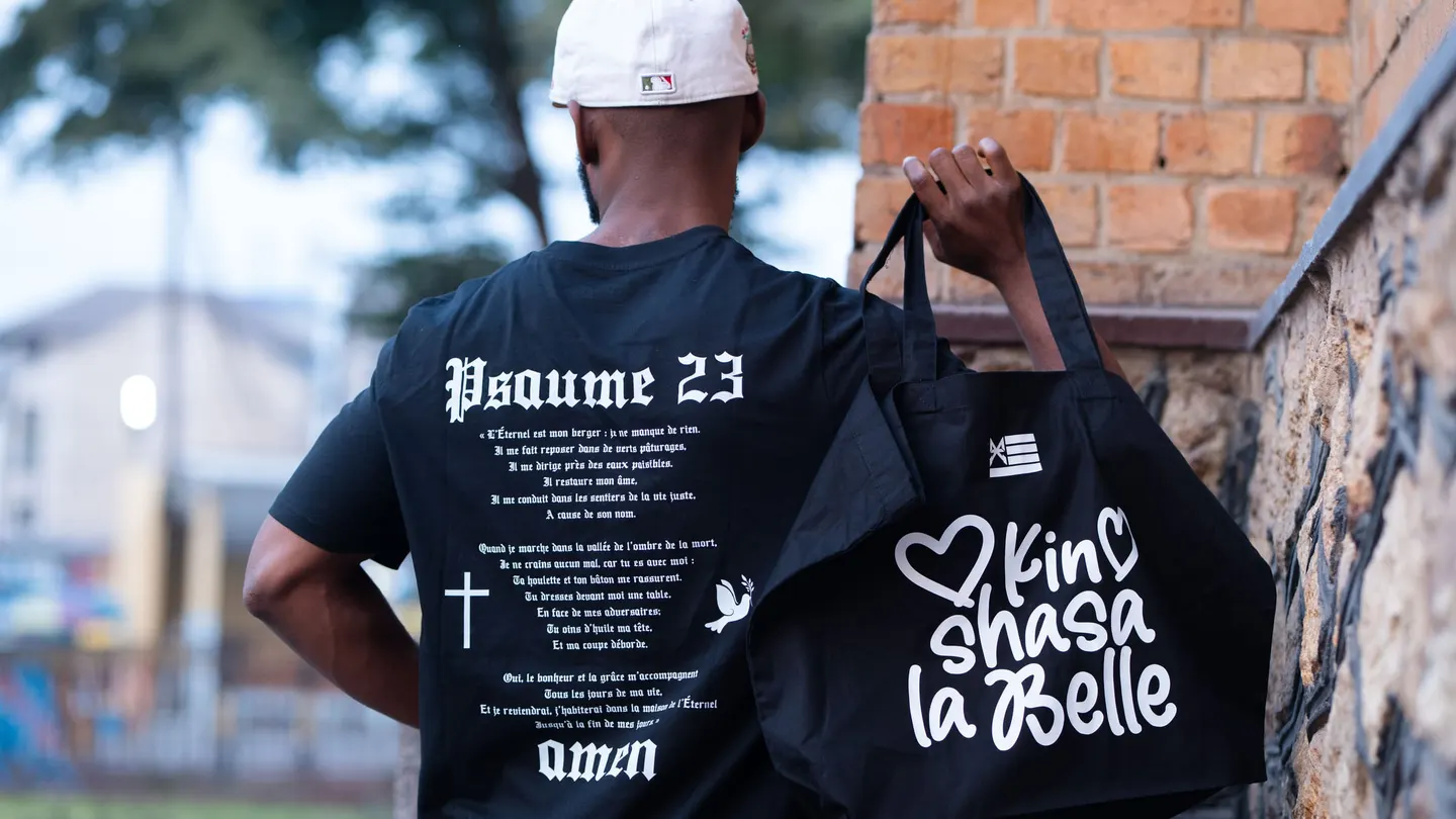 De dos, en t-shirt noir MK Psaume 23, le tote bag « Kinshasa la Belle » à la main, devant un mur de briques à Kinshasa
